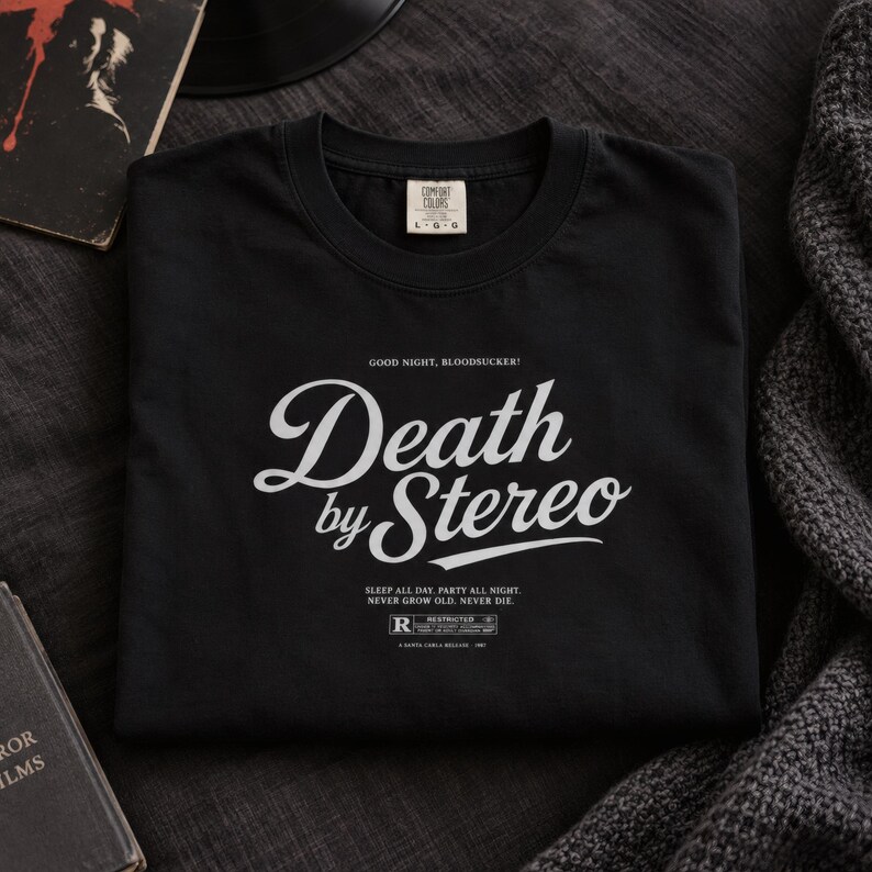 A folded black t-shirt with white Death by Stereo text printed on the chest rests on a dark surface next to a book and blanket.