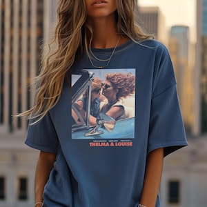 Puede incluir: Una camiseta azul con un estampado gráfico de Thelma y Louise en un coche descapotable. El texto "THELMA & LOUISE" está impreso debajo de la imagen.