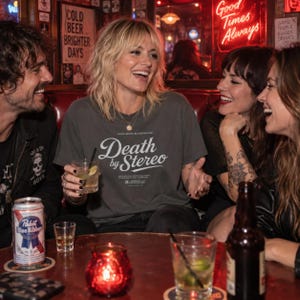 Three friends sit at a dark wooden table in a bar holding drinks, with the center person wearing a dark grey t-shirt that has white Death by Stereo text.