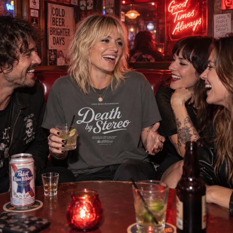 Three friends sit at a dark wooden table in a bar holding drinks, with the center person wearing a dark grey t-shirt that has white Death by Stereo text.