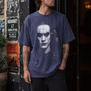 A person with light skin and tattoos wears a dark blue t-shirt with a faded graphic of a pale face with dark eye makeup and red lips.