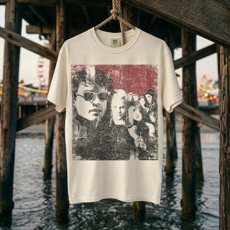 A white t-shirt with a graphic of five people in a red box, hanging on a rope against a dark, watery background under a pier.