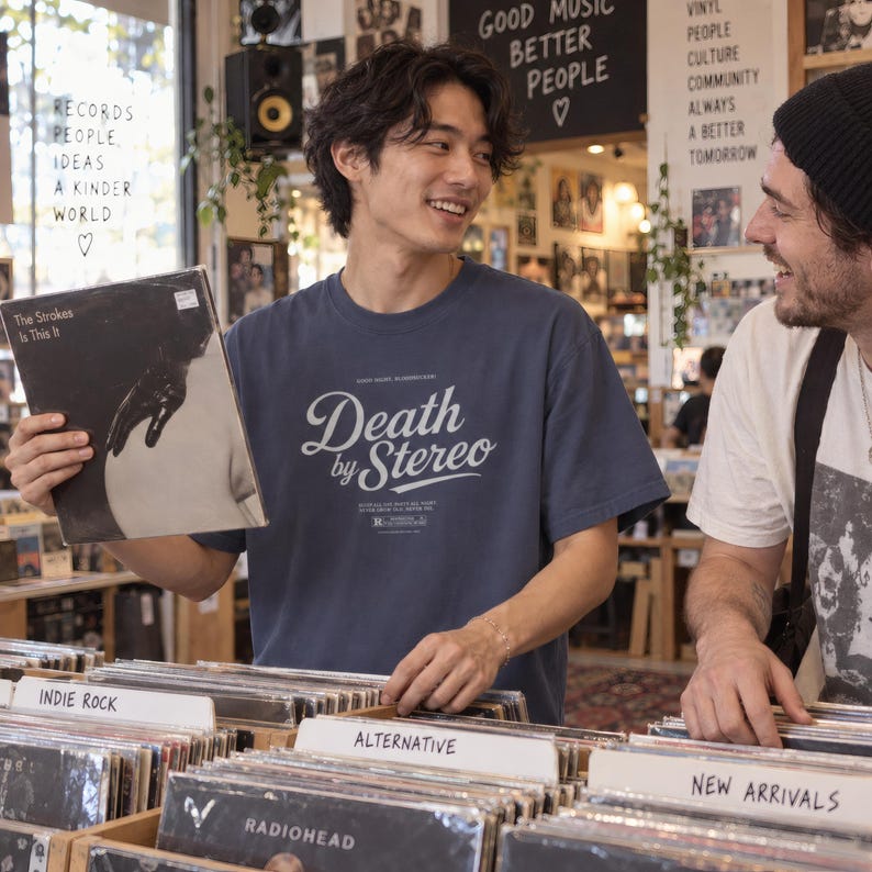 Two people examine vinyl records in a record store, with the person on the left wearing a slate blue t-shirt with white Death by Stereo text.