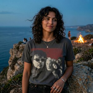 A woman wearing a dark gray t-shirt with a graphic of five people in a red box, sitting on rocks overlooking the ocean at dusk.