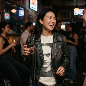A smiling woman in a bar wears a black leather jacket over a white t-shirt with a grayscale portrait of Brandon Lee as The Crow.