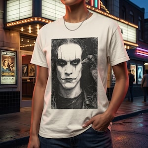 A person stands in front of a movie theater, wearing a white t-shirt with a grayscale portrait of Brandon Lee as The Crow, with a crow on his shoulder.