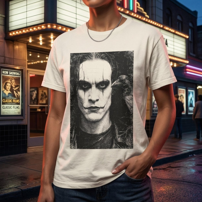 A person stands in front of a movie theater, wearing a white t-shirt with a grayscale portrait of Brandon Lee as The Crow, with a crow on his shoulder.