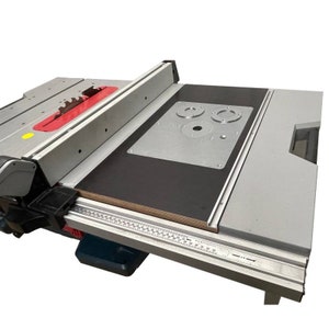 Può includere: Una sega da banco con un piano di lavoro nero e argento, una lama rossa e una piastra di metallo argentata con tre fori. La sega da banco ha una guida in metallo argentata e un righello nero e argento.