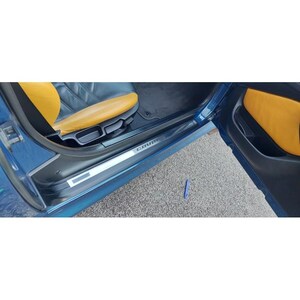 May include: Interior view of a blue coupe car with a yellow and black leather seat. The door is open, revealing a door sill with the word "COUPE" in silver lettering. The car is parked on a gray asphalt surface.