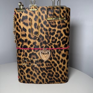 May include: A notebook with a leopard print cover featuring a bright pink elastic band, a gold-toned heart charm, and several gold-toned metal binder clips and paper clips attached to the top and front edges of the cover.
