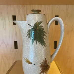 May include: A ceramic coffee pot with a beige glaze and a leaf design in shades of green, brown, and gold. The pot has a rounded body and a curved handle.
