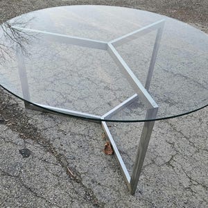Puede incluir: Una mesa de centro moderna con una tapa redonda de cristal y una base de metal cepillado. El cristal transparente refleja el entorno. La base tiene un diseño geométrico único, con tres patas anguladas que se unen en el centro.