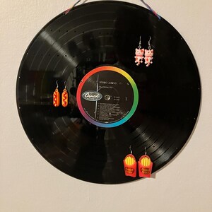 May include: A black vinyl record with a rainbow label is hanging on a wall. Three pairs of earrings are displayed on the record: a pair of hot dog earrings, a pair of pig earrings, and a pair of french fry earrings.