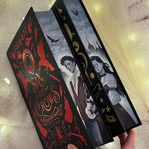 Puede incluir: Una edición de coleccionista de Six of Crows de Leigh Bardugo, con una cubierta negra con detalles en rojo y dorado. El título del libro está en una fuente estilizada y la cubierta presenta un cuervo con un ala roja.