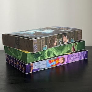 Könnte beinhalten: Ein Stapel von vier dekorativen Boxen mit Illustrationen aus einer Fantasy-Buchreihe. Die Boxen zeigen Charaktere, Szenen und einen Drachen. Die Farben sind lebendig und die Details sind gut dargestellt.