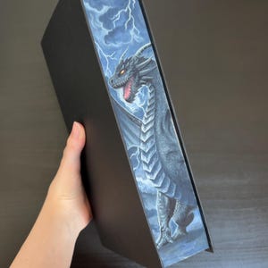 Op de afbeelding: Een zwart boek met een drakenillustratie op de zijkant. De draak is grijs met rode ogen en een open mond, tegen een stormachtige lucht met bliksem. Het boek wordt door een hand vastgehouden.