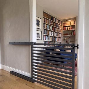 May include: A dark gray metal baby gate with horizontal bars, mounted on wheels. The gate is partially open, revealing a room with a blue sofa and bookshelves filled with books. The gate is installed in a doorway with light wood flooring.
