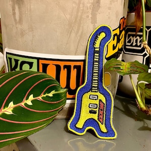 Puede incluir: Un parche de guitarra amarillo y azul con detalles rojos. El parche está bordado y tiene un contorno azul.