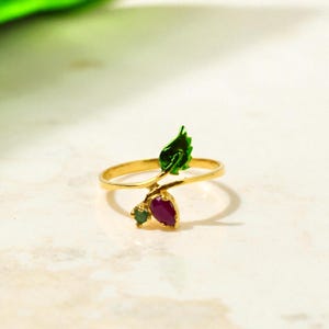 May include: A gold ring with a leaf and berry design. The leaf is green, and the berries are red and green. The ring is set on a white surface.