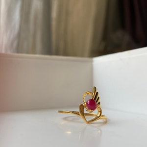 May include: A gold ring featuring a red gemstone set within a heart design with wings.