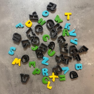 Può includere: Un assortimento di formine per biscotti a forma di alfabeto colorate in nero, giallo, verde e blu. Le formine sono disposte su una superficie grigia, ideali per la cottura e l'artigianato.