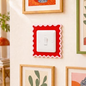 Op de afbeelding: Een witte lichtschakelaar met een rechthoekige frontplaat is geplaatst in een rode, geschulpte lijst. De lijst is gemonteerd op een lichtgekleurde muur, omringd door ingelijste kunstwerken. De schakelaar staat in de 'uit'-stand.