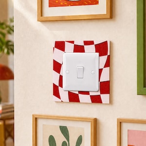 Può includere: Un interruttore bianco con un interruttore rettangolare è montato su una placca decorativa a scacchi rossi e bianchi. La placca è montata su una parete di colore chiaro, con opere d'arte incorniciate visibili sopra e di lato.