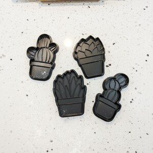Può includere: Quattro formine per biscotti a forma di cactus in plastica nera. Le formine presentano diversi disegni di cactus e piante grasse, tra cui un cactus a botte, una rosetta di succulenta e un cactus in vaso. Le formine sono disposte su una superficie bianca maculata.