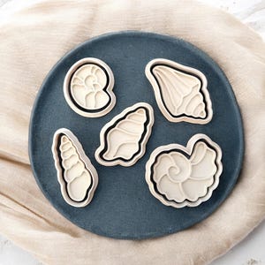 Può includere: Cinque formine per biscotti a forma di conchiglia in vari design, tra cui una spirale e una conchiglia, sono disposte su un piatto blu scuro. Le formine sono bianco sporco con contorni neri.