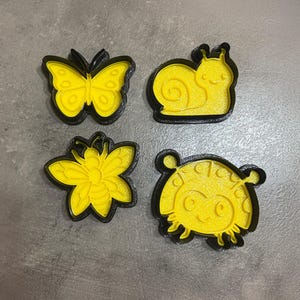 Könnte beinhalten: Vier gelb-schwarze Ausstecher in Insektenform. Die Formen umfassen einen Schmetterling, eine Schnecke, eine Blume und einen Marienkäfer. Die Ausstecher sind aus Kunststoff und zum Backen bestimmt.