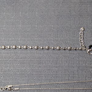 May include: A silver bracelet with round, sparkling accents and a heart-shaped charm with black enamel and clear stones. A silver necklace is also visible, all on a gray background.