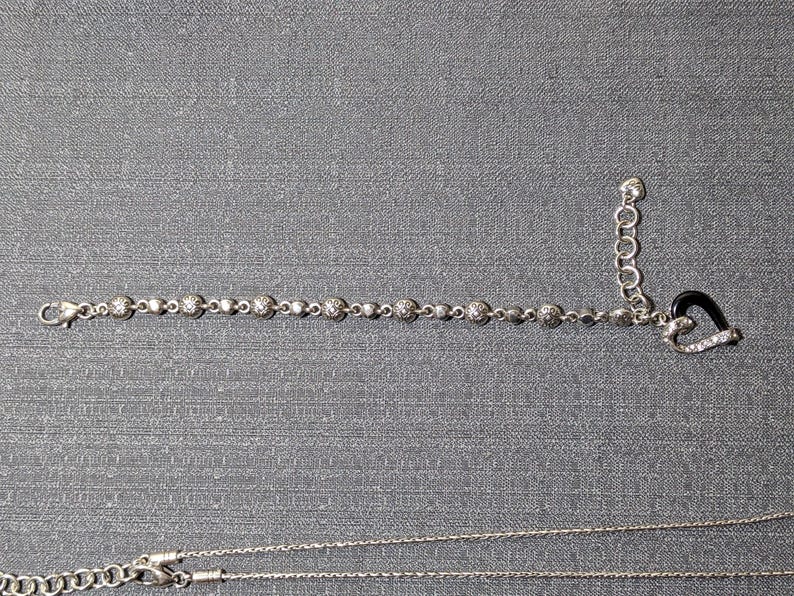 May include: A silver bracelet with round, sparkling accents and a heart-shaped charm with black enamel and clear stones. A silver necklace is also visible, all on a gray background.