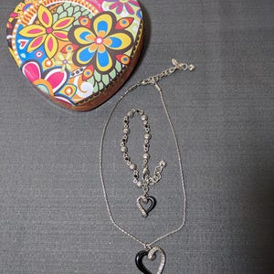 May include: A heart-shaped trinket box with a colorful floral design, a silver necklace with a black heart pendant, and a matching bracelet. The jewelry features clear crystals and black enamel.