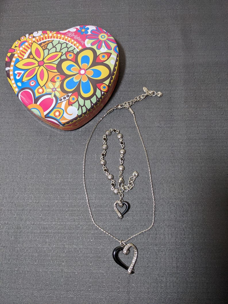 May include: A heart-shaped trinket box with a colorful floral design, a silver necklace with a black heart pendant, and a matching bracelet. The jewelry features clear crystals and black enamel.