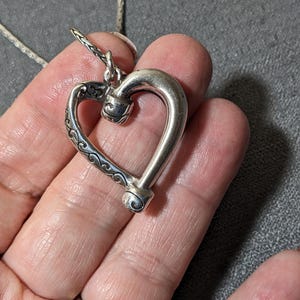 May include: A silver heart pendant necklace. The heart-shaped pendant has a detailed design on one side and a smooth, curved form on the other. The necklace chain is a delicate, silver cable chain.