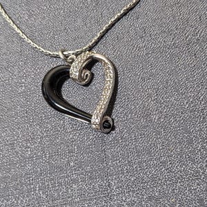 May include: A silver-toned heart pendant necklace. The heart features a black enamel half and a sparkling, rhinestone-studded half. The necklace chain is a delicate, woven design, and the background is a textured gray surface.