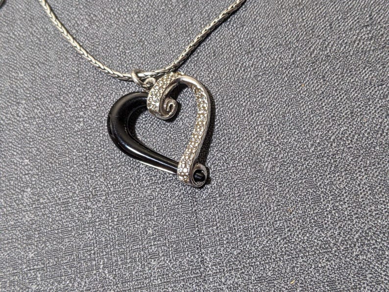 May include: A silver-toned heart pendant necklace. The heart features a black enamel half and a sparkling, rhinestone-studded half. The necklace chain is a delicate, woven design, and the background is a textured gray surface.