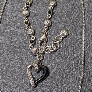 May include: Silver-toned necklace featuring a black heart pendant with a silver-toned, crystal-studded border. The necklace has a decorative chain with black accents and a toggle clasp. The chain is approximately 20 inches long.