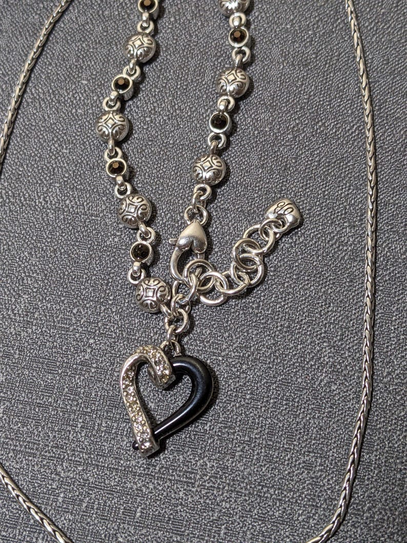 May include: Silver-toned necklace featuring a black heart pendant with a silver-toned, crystal-studded border. The necklace has a decorative chain with black accents and a toggle clasp. The chain is approximately 20 inches long.