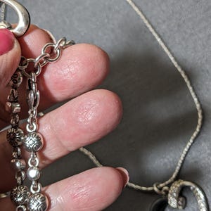 May include: Silver-toned necklace with a decorative pendant and multiple dangling charms. The necklace features a delicate chain and ornate details on the charms. The pendant has a curved design with embedded crystals. The necklace is held by a hand with pink nail polish.