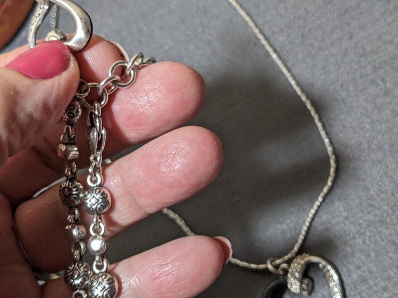 May include: Silver-toned necklace with a decorative pendant and multiple dangling charms. The necklace features a delicate chain and ornate details on the charms. The pendant has a curved design with embedded crystals. The necklace is held by a hand with pink nail polish.