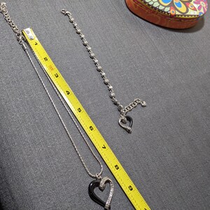 May include: Silver-toned necklace with a black heart pendant, accented with clear crystals. A matching bracelet with a heart charm is also present. A yellow measuring tape is next to the jewelry. A decorative box is in the background.