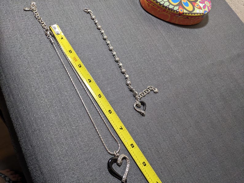 May include: Silver-toned necklace with a black heart pendant, accented with clear crystals. A matching bracelet with a heart charm is also present. A yellow measuring tape is next to the jewelry. A decorative box is in the background.