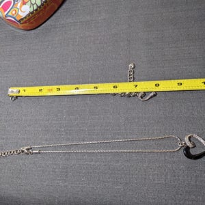 May include: A silver necklace with a black heart-shaped pendant, accented with clear stones. A matching bracelet is also visible. The necklace is approximately 9 inches long. A colorful heart-shaped tin is in the background.
