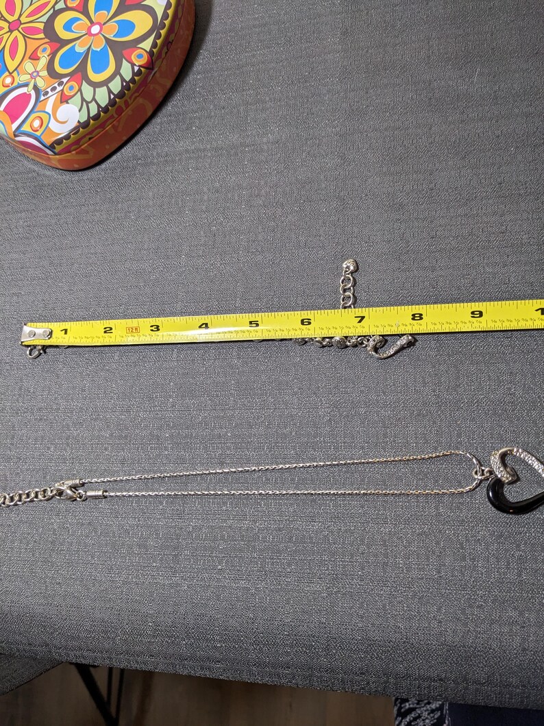 May include: A silver necklace with a black heart-shaped pendant, accented with clear stones. A matching bracelet is also visible. The necklace is approximately 9 inches long. A colorful heart-shaped tin is in the background.