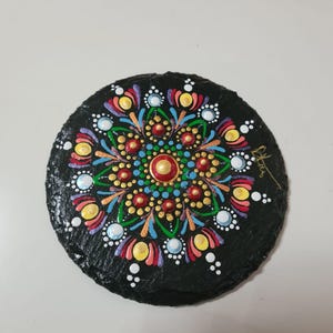 Peut inclure: Un dessous de verre rond et noir, orné d'un mandala peint à la main. Le motif symétrique présente des touches de rouge, jaune, bleu et vert, avec des détails de points blancs et dorés. L'objet est probablement en ardoise.