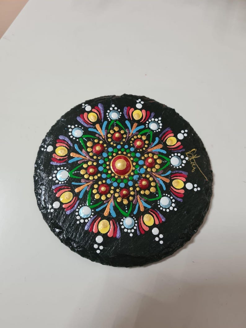 May include: A circular black slate coaster featuring a hand-painted mandala design in vibrant colors including red, yellow, blue, green, and white. The intricate pattern consists of symmetrical dots and petal shapes radiating from a central gold dot.