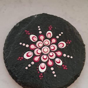 May include: A circular black slate coaster featuring a hand-painted mandala design in the center. The pattern consists of concentric circles and petal shapes created with dots of white, light pink, and dark magenta paint on the textured surface.
