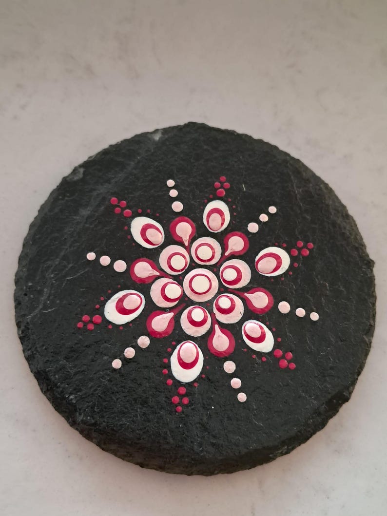 May include: A circular black slate coaster featuring a hand-painted mandala design in the center. The pattern consists of concentric circles and petal shapes created with dots of white, light pink, and dark magenta paint on the textured surface.