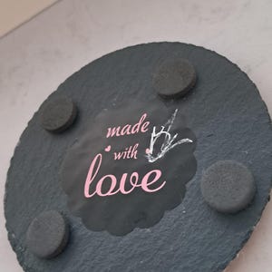 May include: A round, dark gray slate coaster with a textured surface. The center features a black scalloped label with the pink text "made with love" and a small white line-art drawing of a bird. Four circular black foam feet are attached.